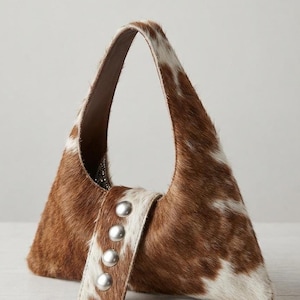 Brown and white cowhide hobo bag with curved shoulder strap and pearl snap button trim, hair-on-hide leather purse for western boho style, shown on a light wood tabletop.