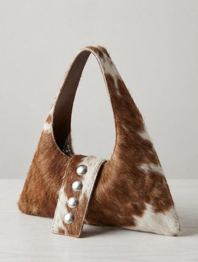 Brown and white cowhide hobo bag with curved shoulder strap and pearl snap button trim, hair-on-hide leather purse for western boho style, shown on a light wood tabletop.