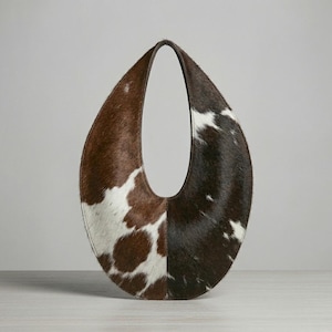 Oval teardrop loop shoulder bag made from genuine tricolor hair-on cowhide, showing brown and dark chocolate panels with natural white markings, photographed standing upright on a grey wooden surface against a light grey wall.