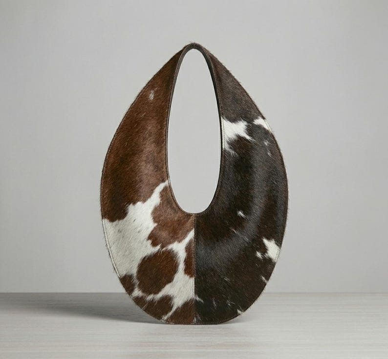 Oval teardrop loop shoulder bag made from genuine tricolor hair-on cowhide, showing brown and dark chocolate panels with natural white markings, photographed standing upright on a grey wooden surface against a light grey wall.