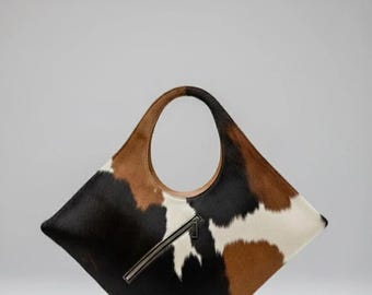 May include: A cowhide tote bag with a distinctive geometric form. The bag displays a blend of black, brown, and white fur, complemented by a brown leather handle and a silver zip. Designed for carrying essentials.