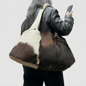 Puede incluir: Un bolso de hombro grande confeccionado en piel animal texturizada con un patrón de bloques de color en pelaje blanco, marrón oscuro y negro. El bolso tiene una correa ancha integrada con un detalle de ojal metálico en el punto de unión superior.