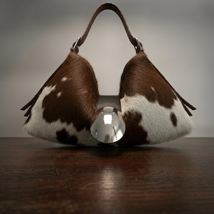 May include: A structured handbag featuring a brown and white cowhide pattern with a distinct triangular silhouette. The bag includes a single brown leather handle and a large, circular, polished silver-tone metal accent centered on the front.