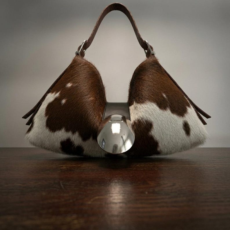 May include: A structured handbag featuring a brown and white cowhide pattern with a distinct triangular silhouette. The bag includes a single brown leather handle and a large, circular, polished silver-tone metal accent centered on the front.