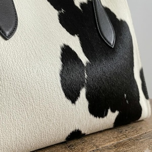 Structured wide-format cowhide tote bag in bold black and white cow print hair-on hide, short dual tapered black leather top handles, open-top trapeze silhouette, self-supporting, photographed front-facing on warm reclaimed wood table against grey wall.