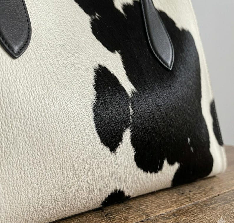 Structured wide-format cowhide tote bag in bold black and white cow print hair-on hide, short dual tapered black leather top handles, open-top trapeze silhouette, self-supporting, photographed front-facing on warm reclaimed wood table against grey wall.