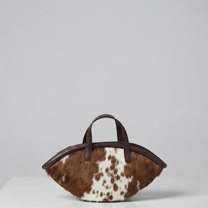 May include: A crescent-shaped handbag featuring a brown and white cowhide pattern with a textured fur finish. The bag includes dark brown leather trim along the edges and two matching dark brown leather handles attached to the top centre.
