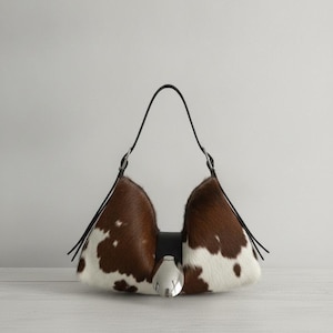 May include: A crescent-shaped handbag featuring a brown and white cowhide pattern with a smooth texture. The bag includes a black leather strap, silver-toned metal hardware, and a prominent silver-colored oval accent centered on the front closure.