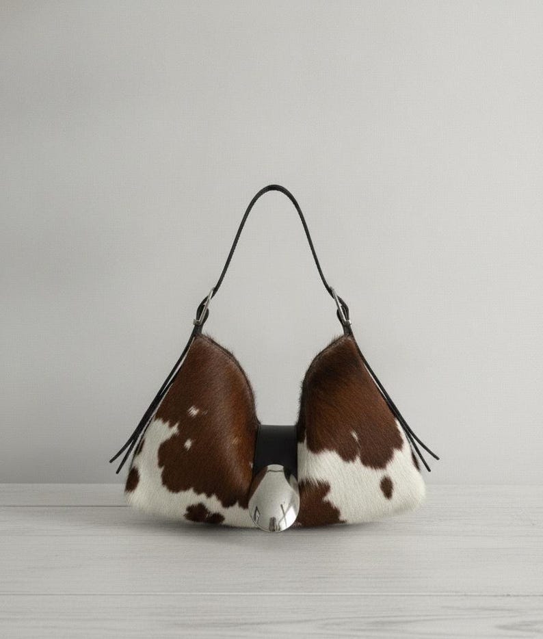 May include: A crescent-shaped handbag featuring a brown and white cowhide pattern with a smooth texture. The bag includes a black leather strap, silver-toned metal hardware, and a prominent silver-colored oval accent centered on the front closure.