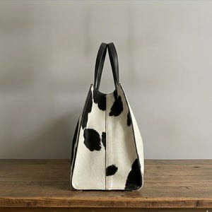 Structured wide-format cowhide tote bag in bold black and white cow print hair-on hide, short dual tapered black leather top handles, open-top trapeze silhouette, self-supporting, photographed front-facing on warm reclaimed wood table against grey wall.