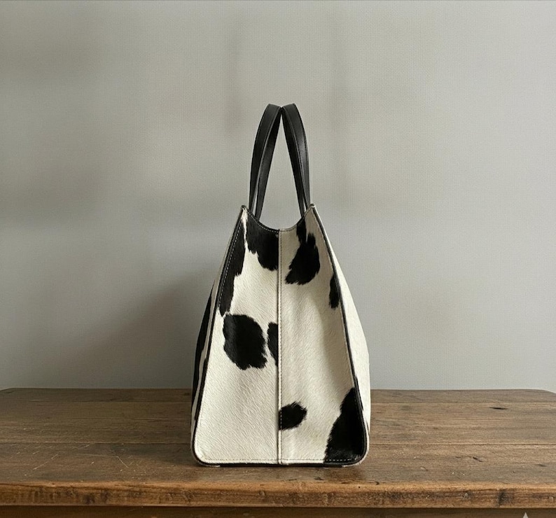 Structured wide-format cowhide tote bag in bold black and white cow print hair-on hide, short dual tapered black leather top handles, open-top trapeze silhouette, self-supporting, photographed front-facing on warm reclaimed wood table against grey wall.
