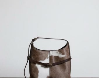 May include: A brown and white cowhide shoulder bag with a curved top and a brown leather strap. The bag has a rectangular shape with a white patch on the front. A smaller brown strap with a buckle crosses the front.
