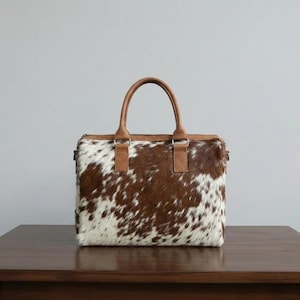 May include: A rectangular handbag featuring a brown and white cowhide pattern with a textured fur finish. The bag includes two tan leather top handles attached with rectangular leather patches and silver-toned metal hardware connectors.