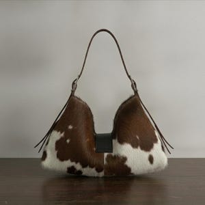 Brown and white speckled cowhide triangular hobo bag with a single dark leather strap, polished silver disc clasp, and fringe tassels, displayed on a dark wood table against a soft grey backdrop.