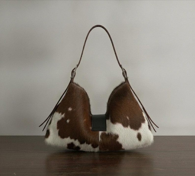 Brown and white speckled cowhide triangular hobo bag with a single dark leather strap, polished silver disc clasp, and fringe tassels, displayed on a dark wood table against a soft grey backdrop.
