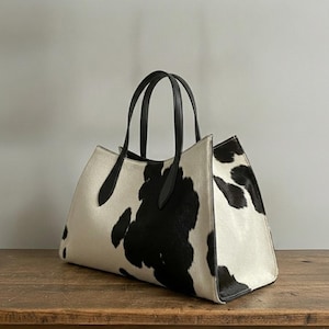 Structured wide-format cowhide tote bag in bold black and white cow print hair-on hide, short dual tapered black leather top handles, open-top trapeze silhouette, self-supporting, photographed front-facing on warm reclaimed wood table against grey wall.