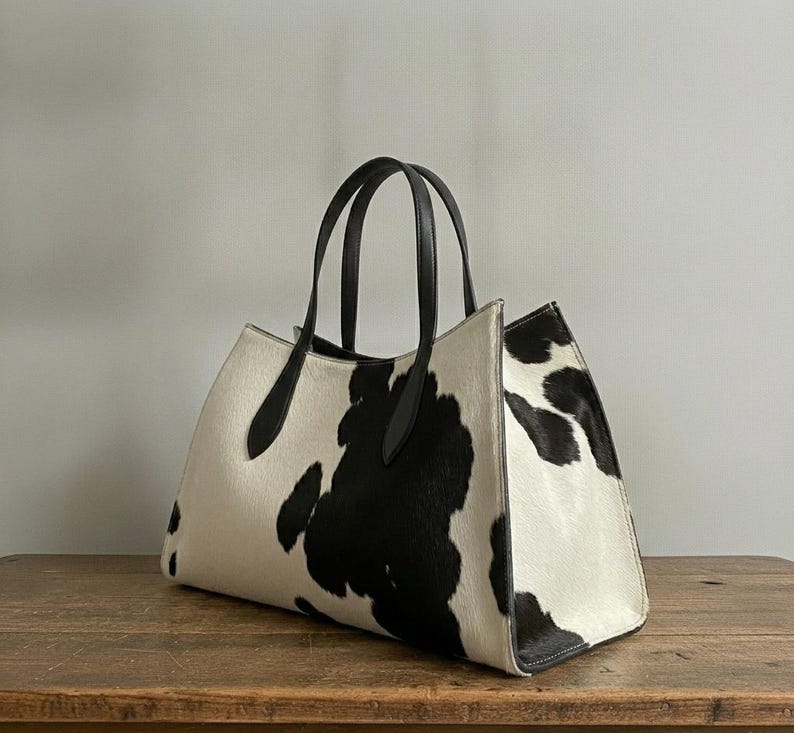 Structured wide-format cowhide tote bag in bold black and white cow print hair-on hide, short dual tapered black leather top handles, open-top trapeze silhouette, self-supporting, photographed front-facing on warm reclaimed wood table against grey wall.