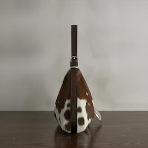 Brown and white speckled cowhide triangular hobo bag with a single dark leather strap, polished silver disc clasp, and fringe tassels, displayed on a dark wood table against a soft grey backdrop.