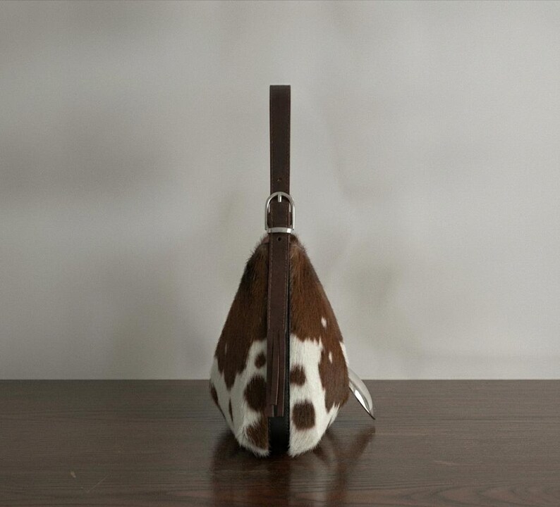 Brown and white speckled cowhide triangular hobo bag with a single dark leather strap, polished silver disc clasp, and fringe tassels, displayed on a dark wood table against a soft grey backdrop.