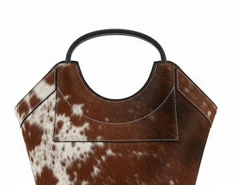May include: A brown and white cowhide tote bag with a black handle. The bag has a unique shape with a curved top and a small pocket on the front. The stitching is white, and the bag is made of a soft, textured material.