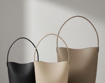May include: Three leather bucket bags in black, beige, and taupe, each with a curved handle. The bags are different sizes, with a textured surface. The bags are displayed on a white marble surface.