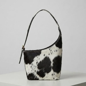 May include: A crescent-shaped shoulder bag featuring a black and white cowhide pattern with a textured fur finish. The bag has a single thin strap attached to one side with a metal buckle detail, creating a distinct asymmetrical silhouette.