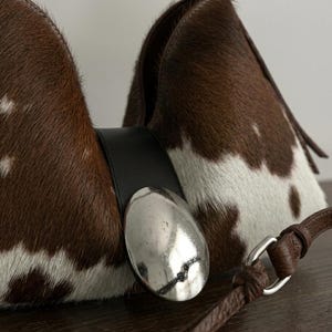 Brown and white speckled cowhide triangular hobo bag with a single dark leather strap, polished silver disc clasp, and fringe tassels, displayed on a dark wood table against a soft grey backdrop.