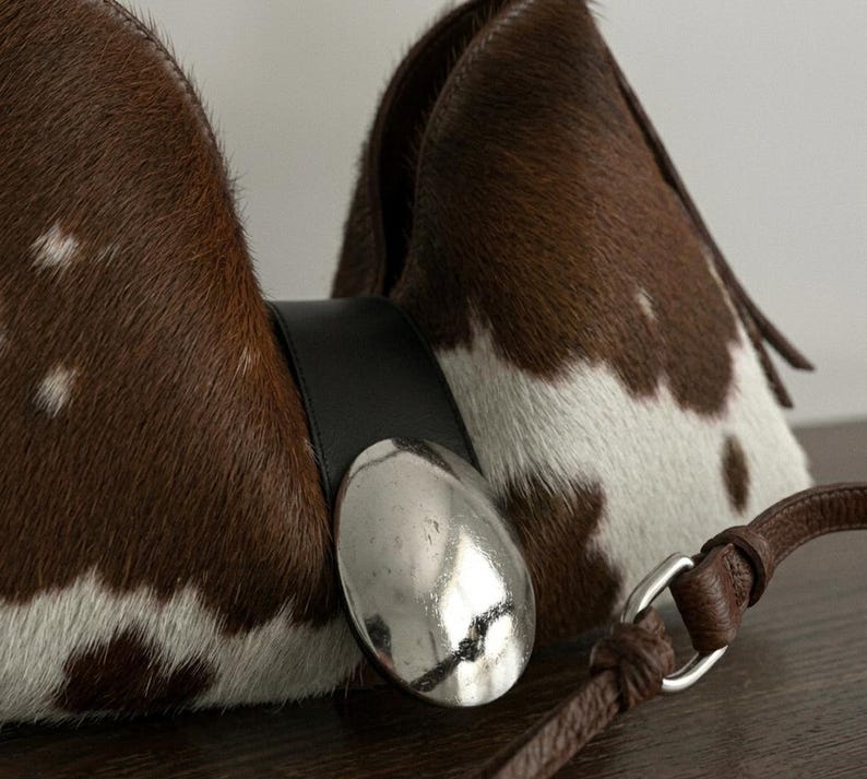 Brown and white speckled cowhide triangular hobo bag with a single dark leather strap, polished silver disc clasp, and fringe tassels, displayed on a dark wood table against a soft grey backdrop.