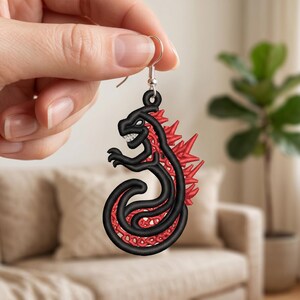 May include: A dangling earring featuring a black embroidered outline of a stylized monster with a curled tail and sharp red spikes along its back. The design is filled with a red lattice pattern and hangs from a silver-toned metal hook.