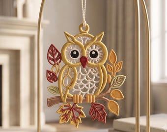 May include: A hanging owl ornament crafted from intricate yellow, white, and red embroidery thread. The owl is perched on a brown branch surrounded by autumn-colored leaves in shades of red, orange, and green, suspended by a white string.