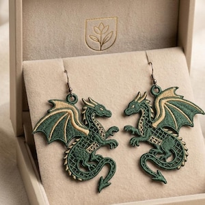 Könnte beinhalten: Ein Paar Ohrhänger mit gestickten grünen und goldenen Drachenmotiven. Jeder Drache ist im Profil mit detaillierten Flügelmustern und einem langen Schwanz dargestellt und an dunklen Metallhaken befestigt. Die Ohrringe liegen in einer beigen Box.