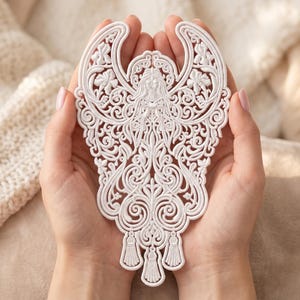 May include: A white, intricately designed angel ornament with detailed wings, flowing hair, and decorative patterns. The angel has a detailed face and three tassel-like elements at the bottom. The ornament is held in cupped hands.