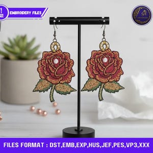 Könnte beinhalten: Ein Paar bestickte Rosenohrringe in Rot-, Gold- und Grüntönen, präsentiert auf einem schwarzen Ständer. Die Ohrringe weisen detaillierte Blumendesigns mit einem perlenartigen Zentrum auf. Der Text "EMBROIDERY FILES" und "FILES FORMAT: DST, EMB, EXP, HUS, JEF, PES, VP3,XXX" ist sichtbar.