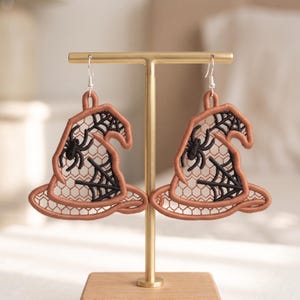 May include: A pair of dangling earrings shaped like witch hats, featuring a copper-colored embroidered outline and a delicate tan lace mesh interior. Each hat is decorated with a black embroidered spider and a black spiderweb pattern.