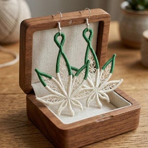 May include: Embroidered leaf earrings in a wooden box. The earrings have a green and white design with a detailed, openwork pattern. They hang from silver-coloured hooks. The box is lined with a neutral fabric, and is displayed on a wooden surface.