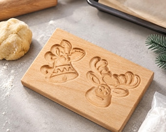 May include: A rectangular light-colored wooden cookie mold featuring two carved designs: a bell with a bow and a reindeer head with antlers. The mold has a smooth, flat surface and is designed for pressing patterns into dough for baking cookies.