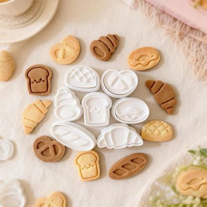 Puede incluir: Una variedad de galletas horneadas y cortadores de galletas. Las galletas incluyen cruasanes, pan, pretzels y panecillos. Los cortadores de galletas son blancos y tienen forma de galletas. Las galletas son de color marrón claro y beige.