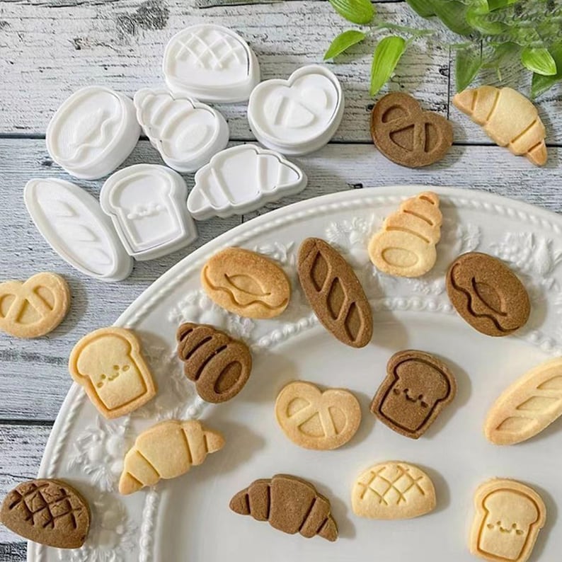 May include: A collection of bread-shaped cookie cutters and baked cookies. The white plastic cutters are in various shapes, including a croissant, pretzel, and loaf of bread. The baked cookies are light and dark brown, with detailed designs.
