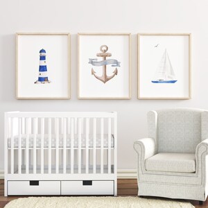May include: Three framed watercolor prints depicting nautical themes: a lighthouse, an anchor with a ribbon, and a sailboat. The prints are displayed on a white wall above a white crib and a white armchair.