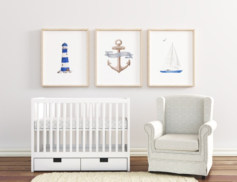 May include: Three framed watercolor prints depicting nautical themes: a lighthouse, an anchor with a ribbon, and a sailboat. The prints are displayed on a white wall above a white crib and a white armchair.