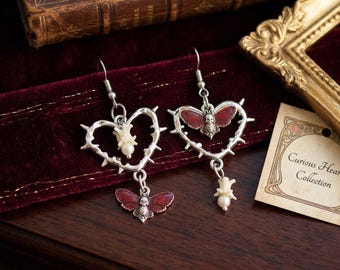 May include: Silver-toned heart-shaped earrings with thorn accents, featuring a red butterfly design and a small skull. Each earring has a dangling pearl and a tag that reads "Curious Hearts Collection."