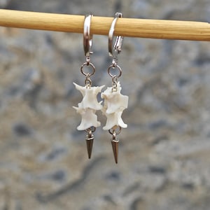 May include: A pair of unique earrings featuring white bone-like pendants with silver-colored metal accents. The earrings are suspended from silver-colored hoops and have a spike-shaped charm at the bottom. The earrings are displayed against a blurred background.