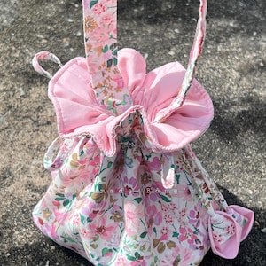 May include: A pink floral drawstring bag with a matching strap and ruffled top. The bag features a delicate pattern of pink and white flowers with green leaves. The bag is made of fabric.