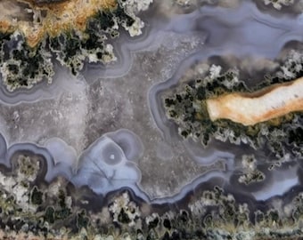 May include: Close-up of a polished stone with a natural, abstract pattern. The stone features layers of gray, white, brown, and green hues, creating a swirling, organic design. The texture appears smooth with visible variations in color and pattern.