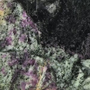May include: Close-up of a natural stone surface with a mix of colours. The stone features a speckled pattern with shades of green, black, and purple. The texture appears rough and uneven, with variations in colour and density.