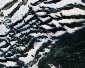May include: Abstract close-up of a stone surface with a unique pattern. The design features a blend of white, black, and dark green, creating a zebra-like effect. The natural stone has a polished appearance, with a mix of smooth and textured areas.