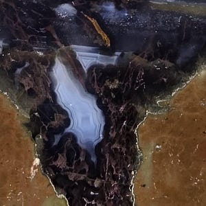 May include: Close-up of a polished stone with a complex pattern. The stone features a mix of colors, including brown, black, and blue, with intricate veining and a polished surface. The overall effect is a natural, organic design.