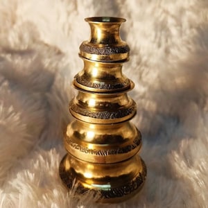 Puede incluir: Un jarrón apilado de color dorado con un diseño detallado y texturizado alrededor de cada sección. El jarrón tiene una parte superior acampanada y una serie de secciones redondeadas y bulbosas. El jarrón decorativo es probablemente de metal y tiene una estética antigua o vintage.
