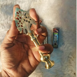 Puede incluir: Un porta incienso de metal color dorado con un diseño en forma de árbol en la parte superior. El soporte tiene una base decorativa y un tallo largo. El porta incienso se sostiene en una mano.