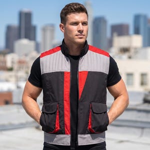May include: A black, red, and gray vest with two front pockets. The vest has a quilted gray upper section and red vertical stripes. The model is wearing a black t-shirt. The background features a cityscape under a bright blue sky.